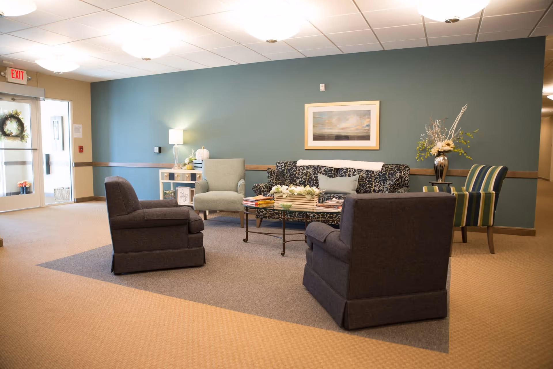 A cozy seating area in a senior living facility with a patterned sofa, two dark armchairs, a light green armchair, and a striped armchair arranged around a glass coffee table. The walls are painted teal with a framed landscape painting, and there is a small white cabinet with a lamp and decorative items. The area is well-lit with ceiling lights and has a beige carpet with a gray rug under the seating.