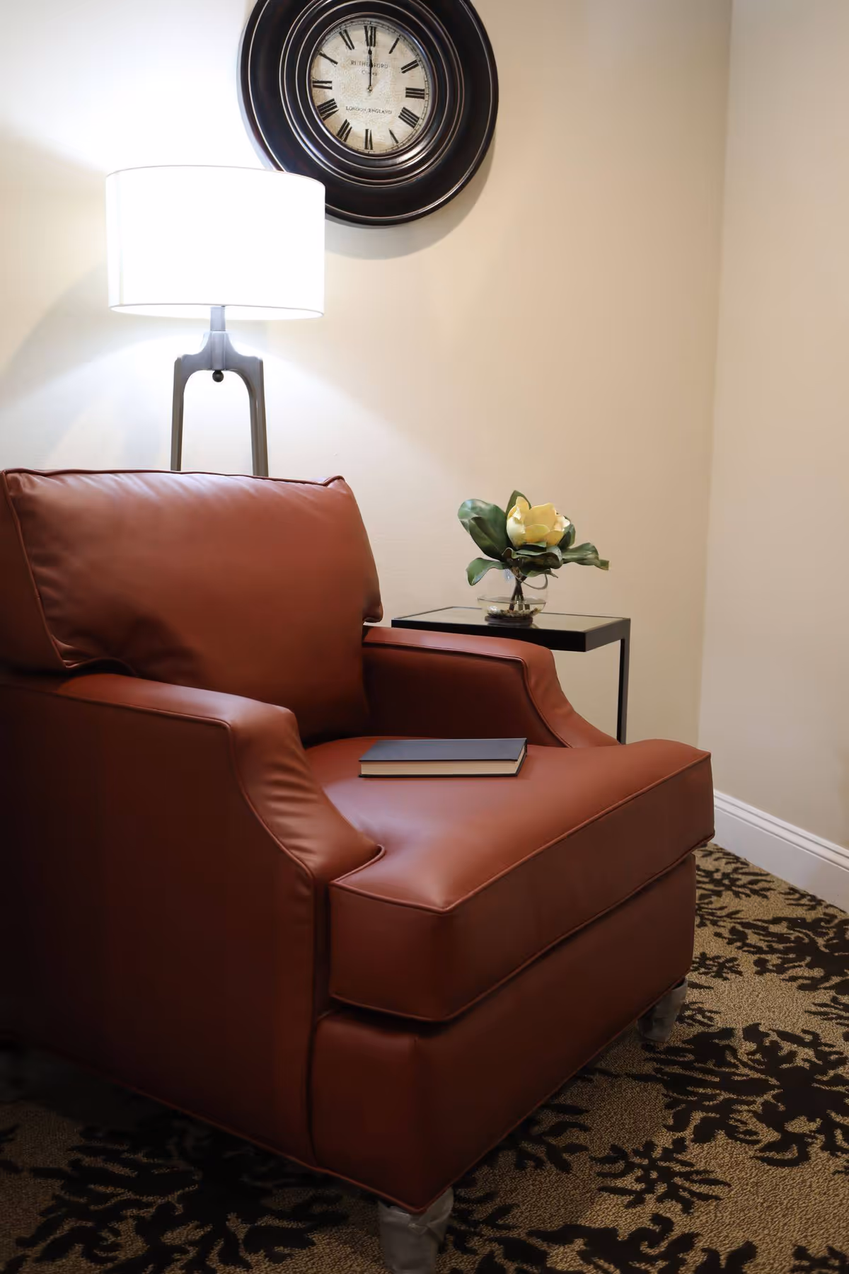 A cozy corner with a brown leather armchair, a closed book resting on the seat, a small side table with a glass vase holding a white flower, a floor lamp with a white shade, and a round wall clock showing the time as 11:55.
