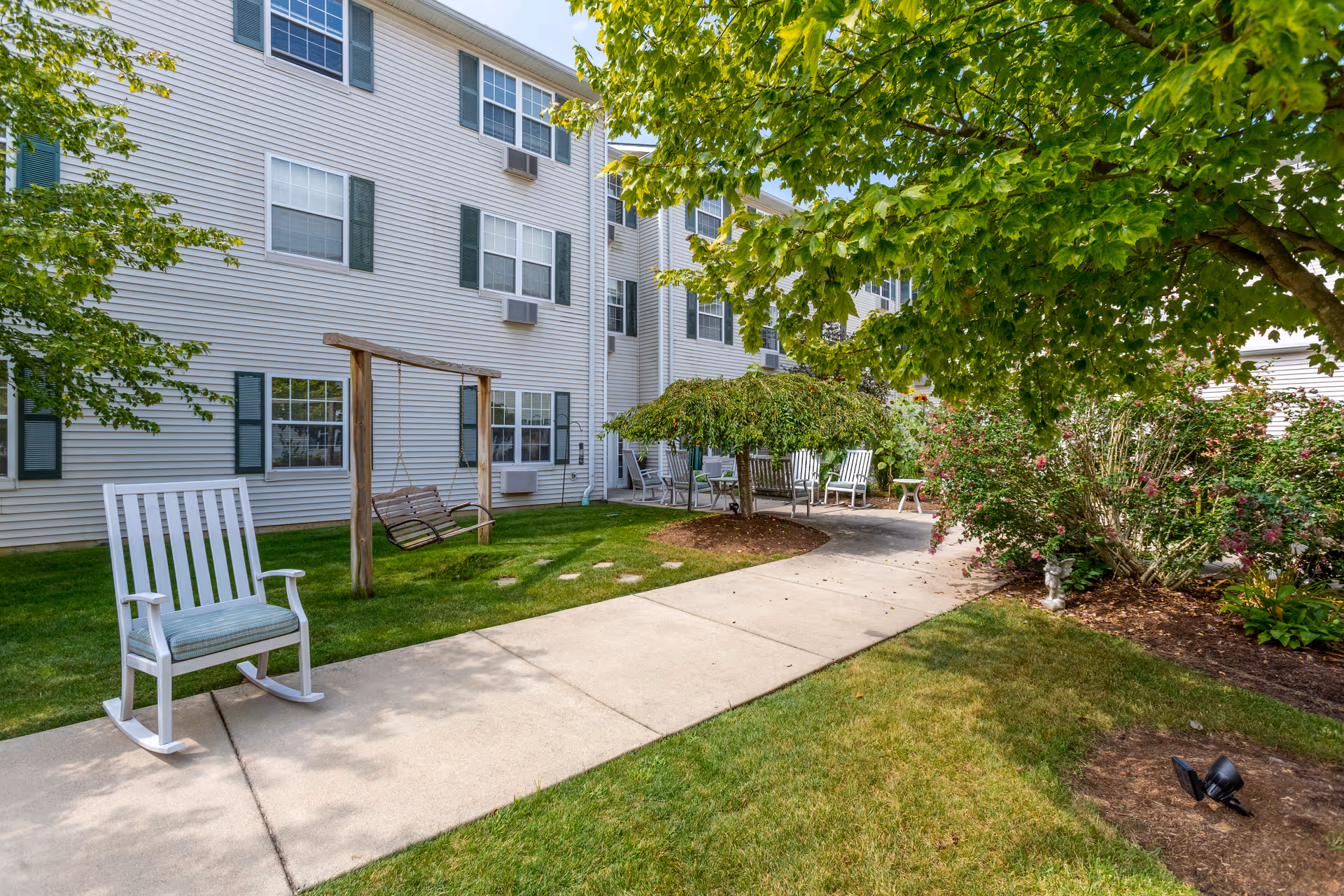 Outdoor garden area at Independence Village of Fishers South featuring a concrete walkway, white rocking chairs, a wooden swing, green grass, trees, and flowering bushes next to a multi-story building with white siding and green shutters.