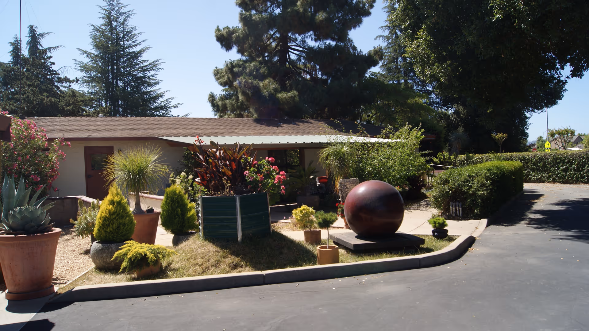 Single-story building entrance with potted plants, shrubs, a large spherical sculpture, and tall trees in the background.