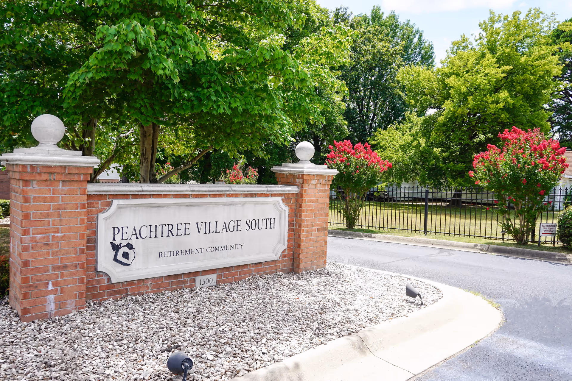 Brick entrance sign that reads 'Peachtree Village South Retirement Community' with trees, pink flowering bushes, and a fenced lawn behind it.