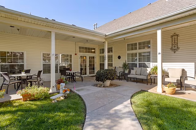 Covered courtyard patio with chairs, tables, potted plants and a walkway leading to glass doors of the senior living facility.