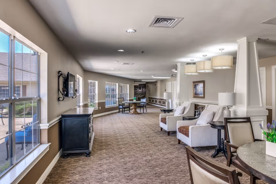 A bright and spacious senior living facility common area with large windows on the left letting in natural light. The room features comfortable armchairs with side tables and lamps, a round table with chairs, and neutral-colored walls and carpet. Decorative lighting fixtures hang from the ceiling, and there is a framed picture on the wall.