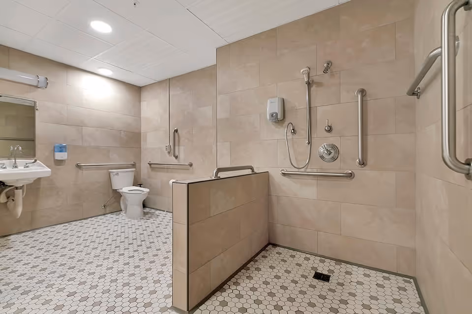Spacious accessible restroom with a toilet, wall-mounted sink, tiled walls and floor, and a roll-in shower with grab bars.