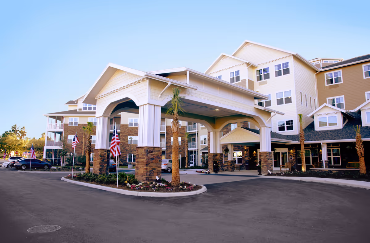 Exterior view of Cypress Springs Gracious Retirement Living facility showing a large covered entrance with stone pillars, palm trees, American flags, and a multi-story building with many windows under a clear blue sky.