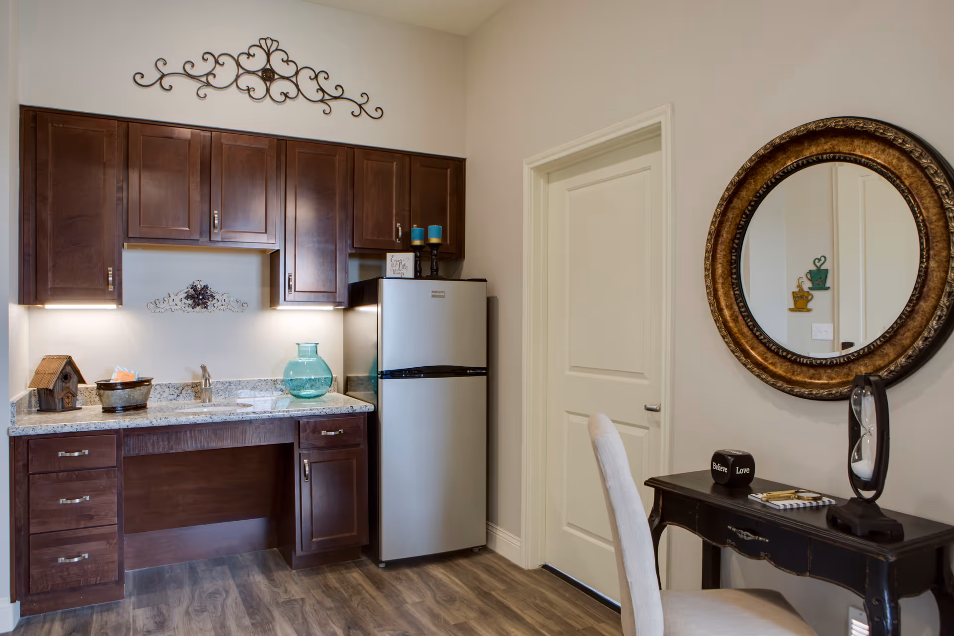 A small kitchen area with dark wooden cabinets, a granite countertop, a stainless steel refrigerator, and decorative items including a birdhouse, a glass vase, and candles. To the right, there is a black wooden desk with a beige chair, a round ornate mirror on the wall, and decorative objects on the desk including an hourglass and a small block with the words 'Believe' and 'Love'.