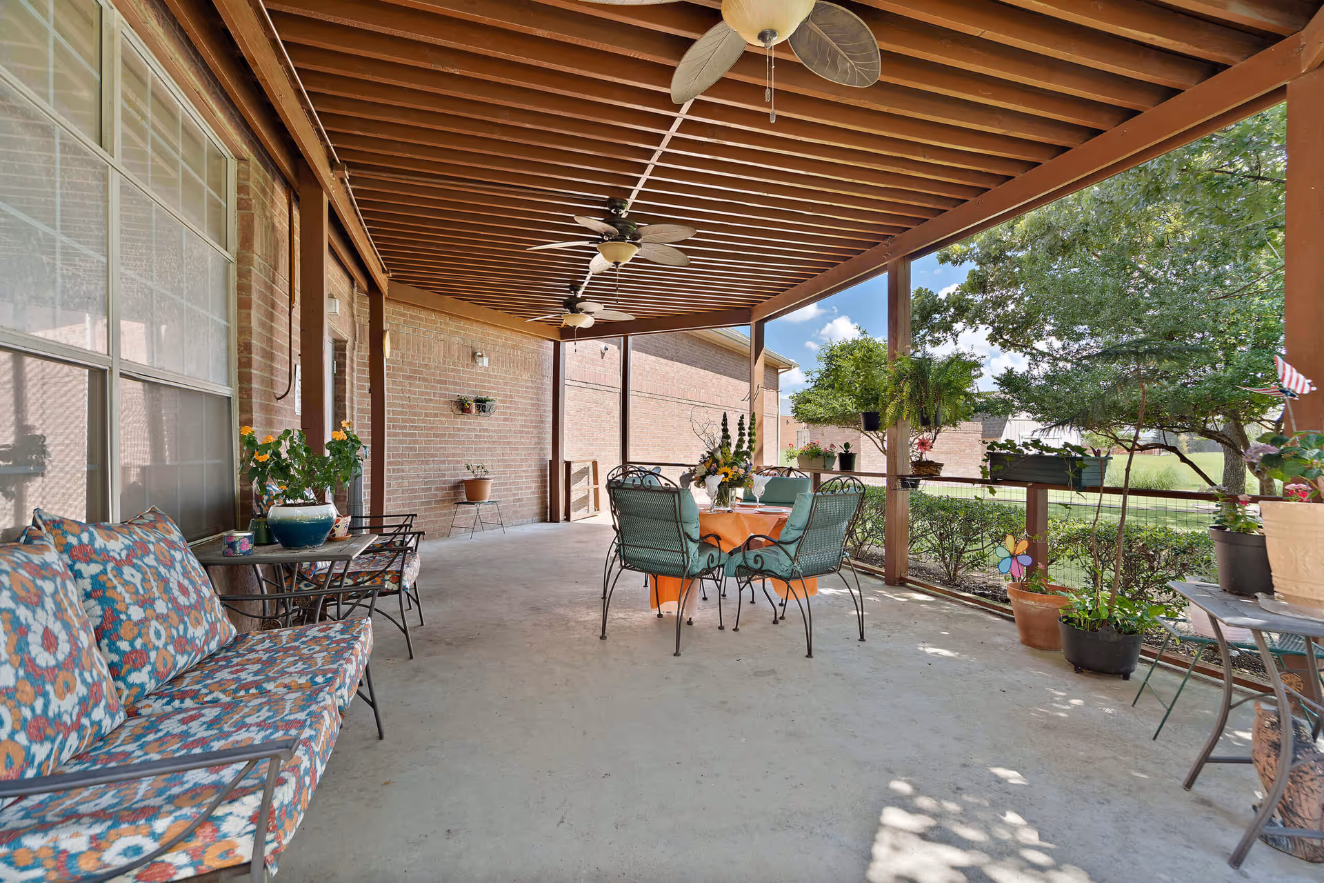 Covered outdoor patio area with a wooden slatted roof, ceiling fans, and a concrete floor. There is a floral cushioned bench and a small table with chairs on the left side, and a round table with four cushioned chairs in the center. Various potted plants and greenery are placed around the patio, with a view of trees and bushes beyond the railing.