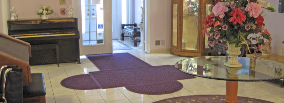 Lobby interior showing a piano, glass entrance doors, purple rugs, a round glass table with a large flower arrangement, and a display cabinet.