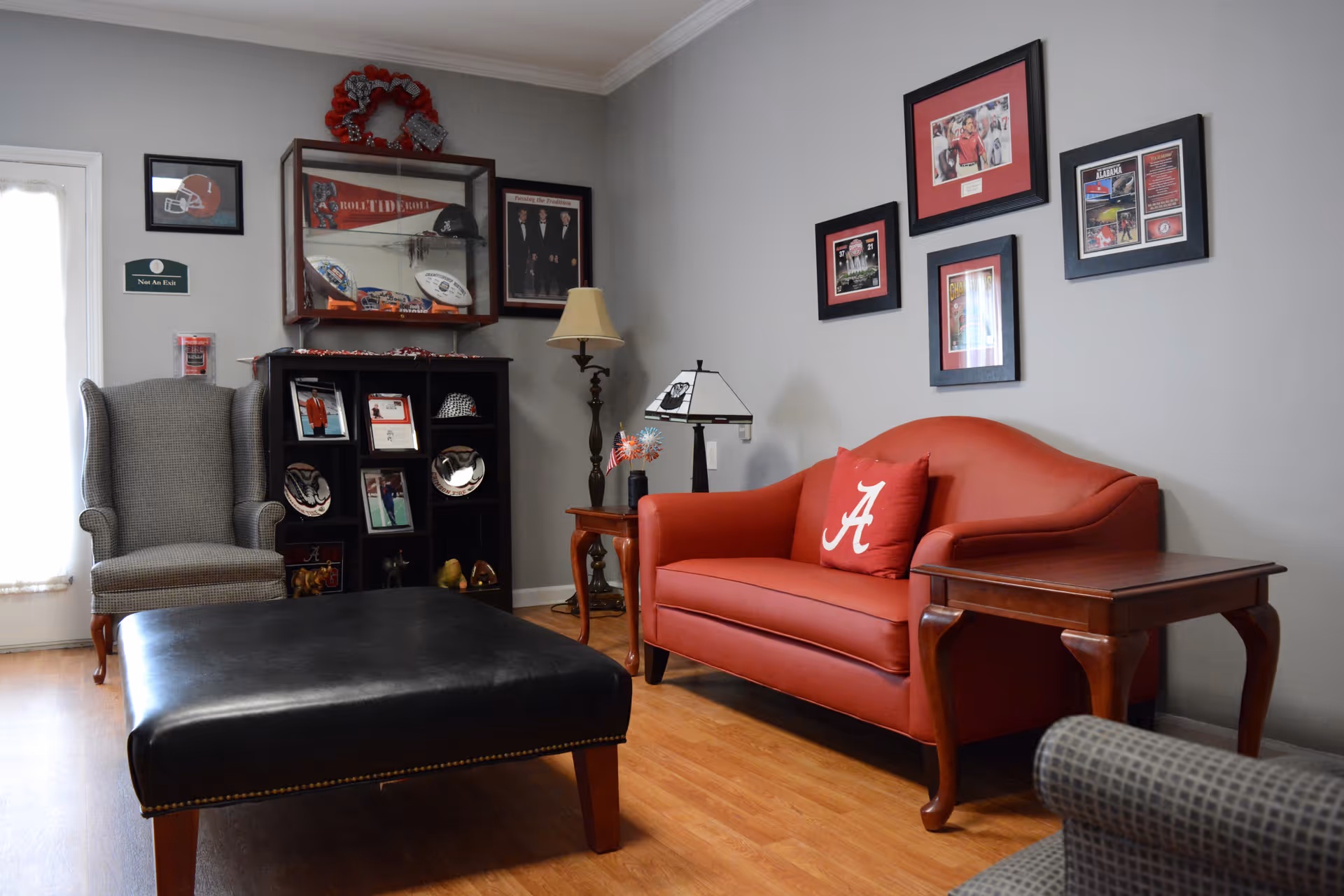 A cozy living room area with a red loveseat featuring a pillow with a white 'A' on it, a black leather ottoman, a gray armchair, wooden side tables, and a display cabinet filled with memorabilia and framed pictures on the walls.