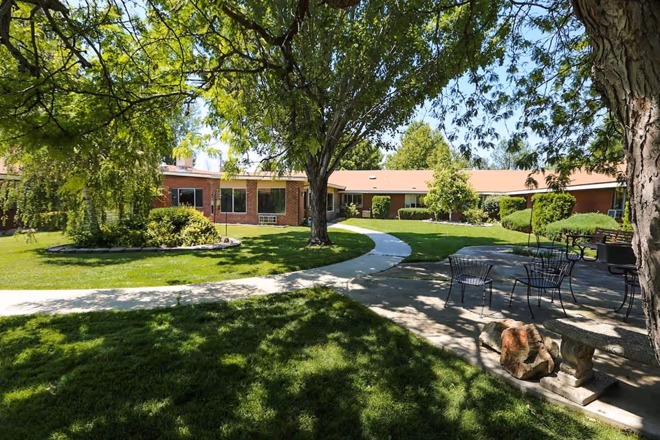 A sunny outdoor courtyard area at Heritage Assisted Living of Boise featuring a curved concrete walkway, green grass, mature trees providing shade, and a patio with metal chairs and a table. The single-story brick building surrounds the courtyard with windows facing the garden.