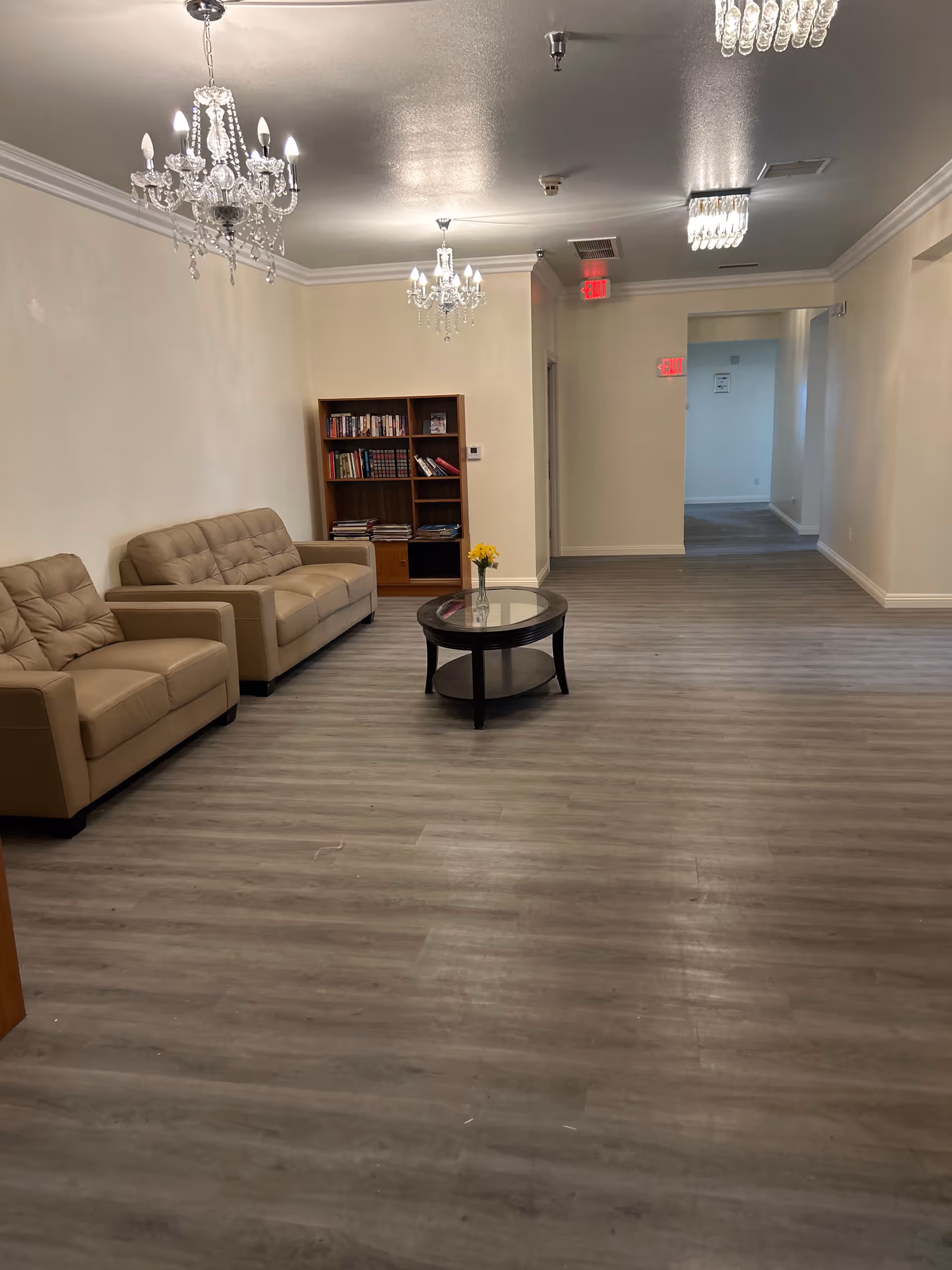 A spacious living room area with two beige leather sofas along the left wall, a round glass-top coffee table with a small vase of yellow flowers in the center, and a wooden bookshelf filled with books against the back wall. The room has light-colored walls, gray wood flooring, and multiple crystal chandeliers hanging from the ceiling. There are exit signs visible in the hallway leading out of the room.