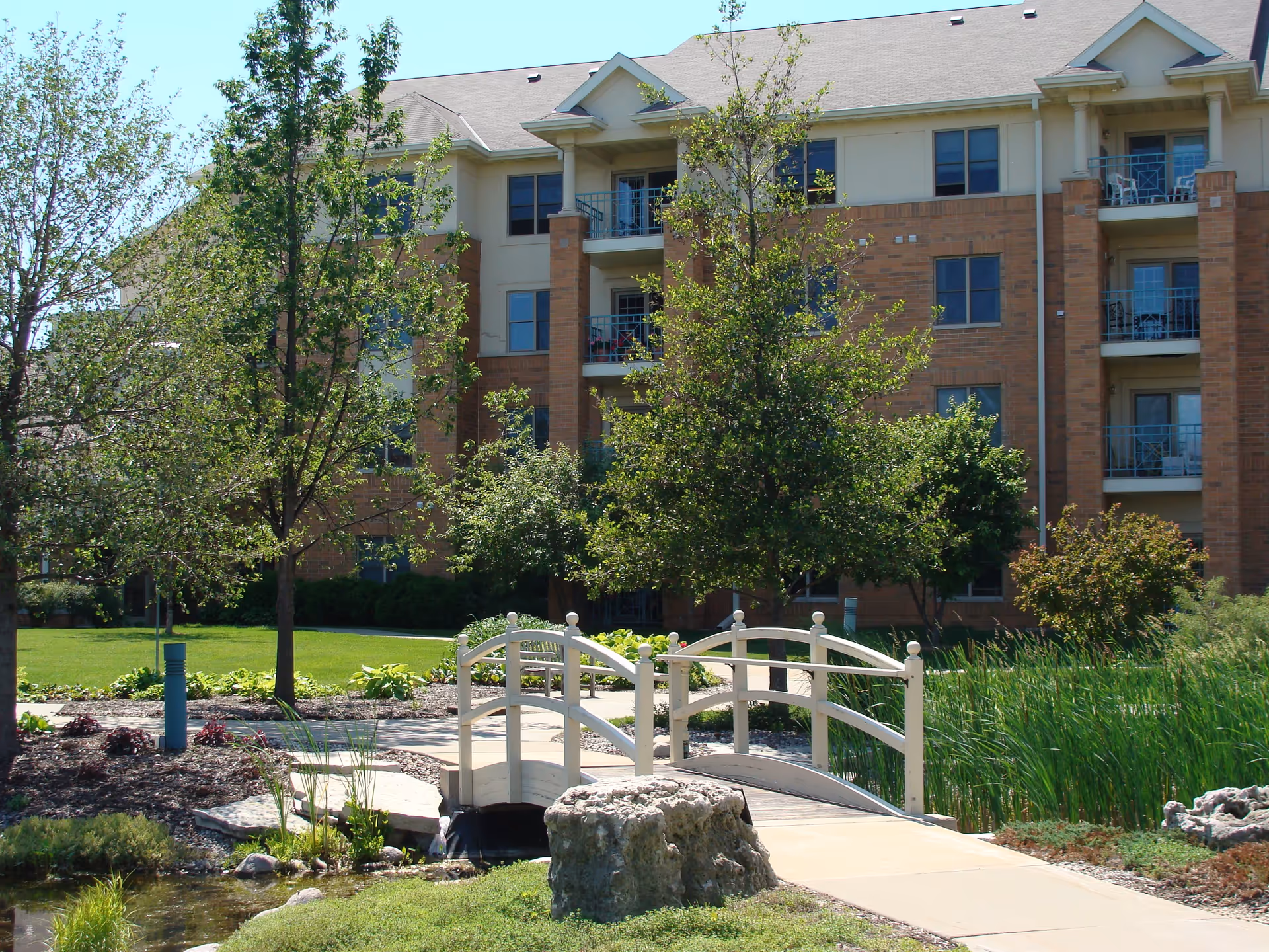 Front of a multi-story brick senior living building with trees, a small decorative bridge, and a pond in the landscaped courtyard.