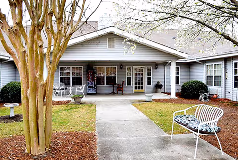 Covered front porch of a single-story building with a concrete walkway, outdoor benches and chairs, trees, and potted plants.