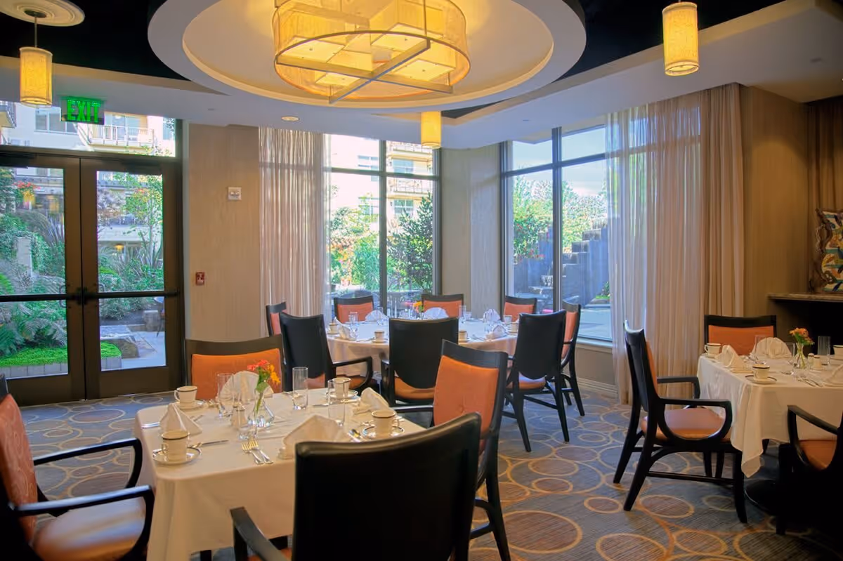 A dining room with round tables covered in white tablecloths, set with cups, glasses, napkins, and silverware. The room has large windows with sheer curtains allowing natural light to enter, and there are cushioned chairs with orange seats around the tables. A large decorative ceiling light fixture and smaller pendant lights hang from the ceiling. Outside the windows, greenery and a garden area are visible.