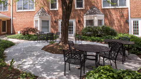 Outdoor courtyard area with round metal tables and chairs arranged on a curved concrete pathway surrounded by greenery and bushes. The background features a brick building with multiple windows and doors, and a large tree in the center of the courtyard.