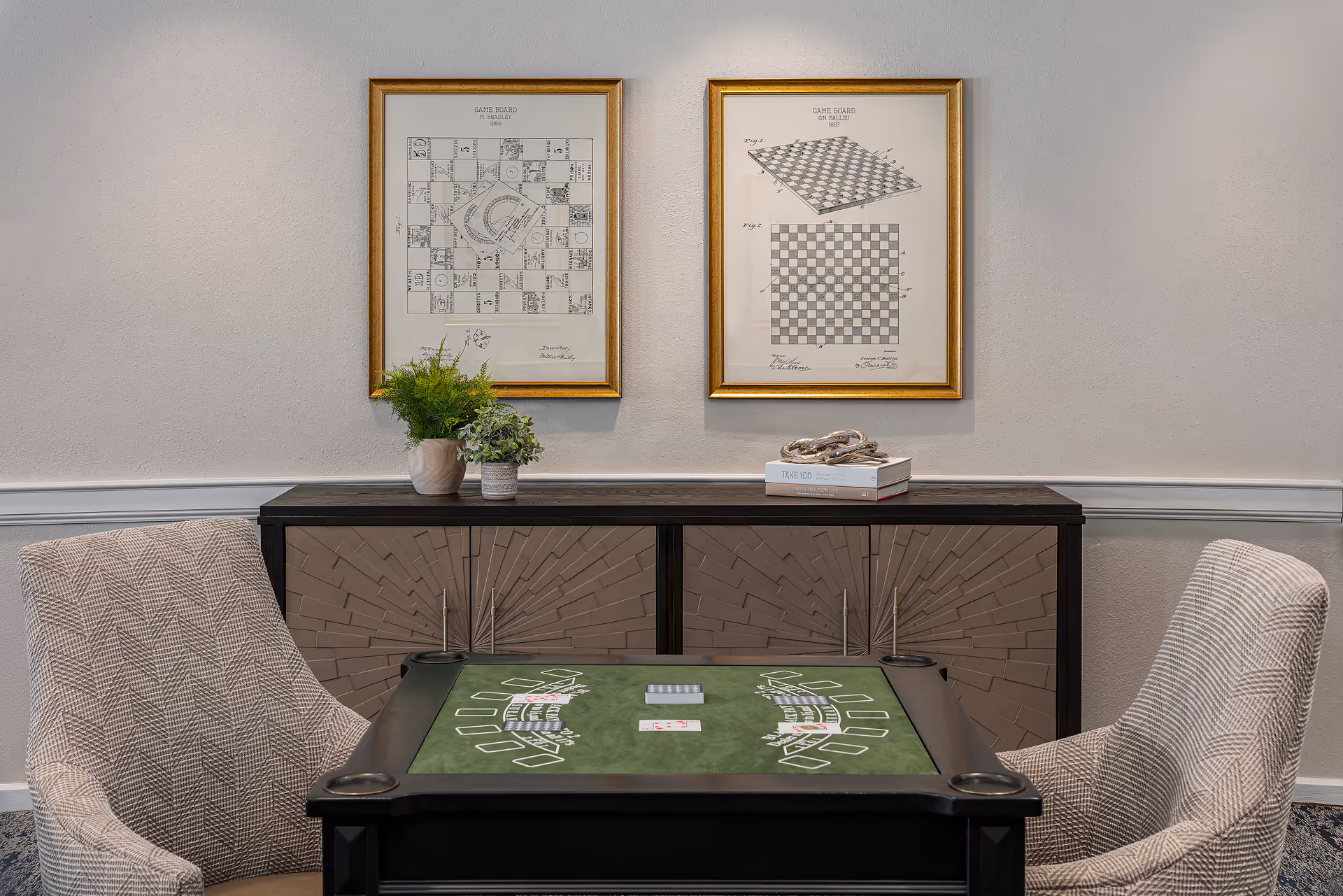A small game table with a green felt surface and card game layout, flanked by two patterned upholstered chairs. Behind the table is a decorative cabinet with two potted plants and a stack of books on top. Above the cabinet, two framed vintage game board diagrams hang on a light gray wall.