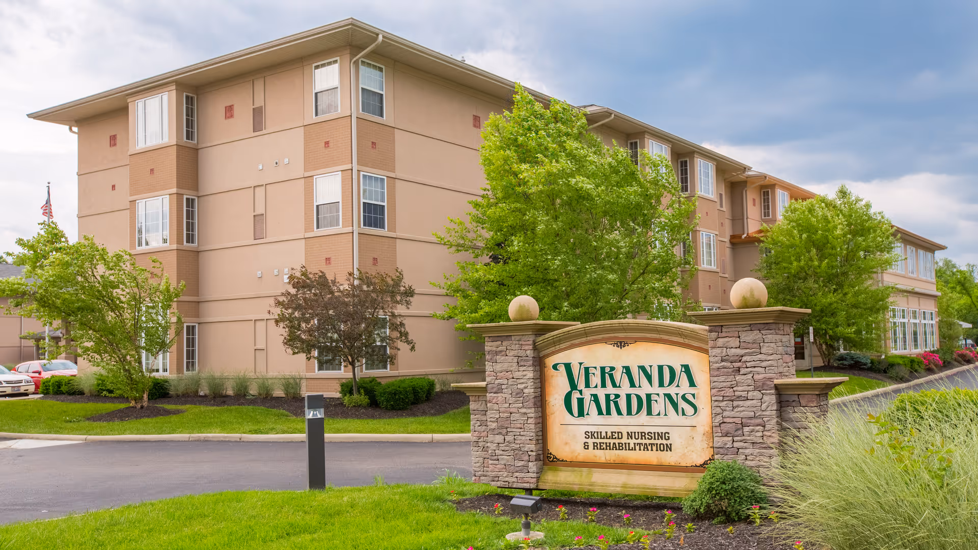 Exterior view of a multi-story beige building with several windows, surrounded by green trees and landscaping. In front of the building is a stone sign that reads 'Veranda Gardens Skilled Nursing & Rehabilitation'. The sky is partly cloudy.