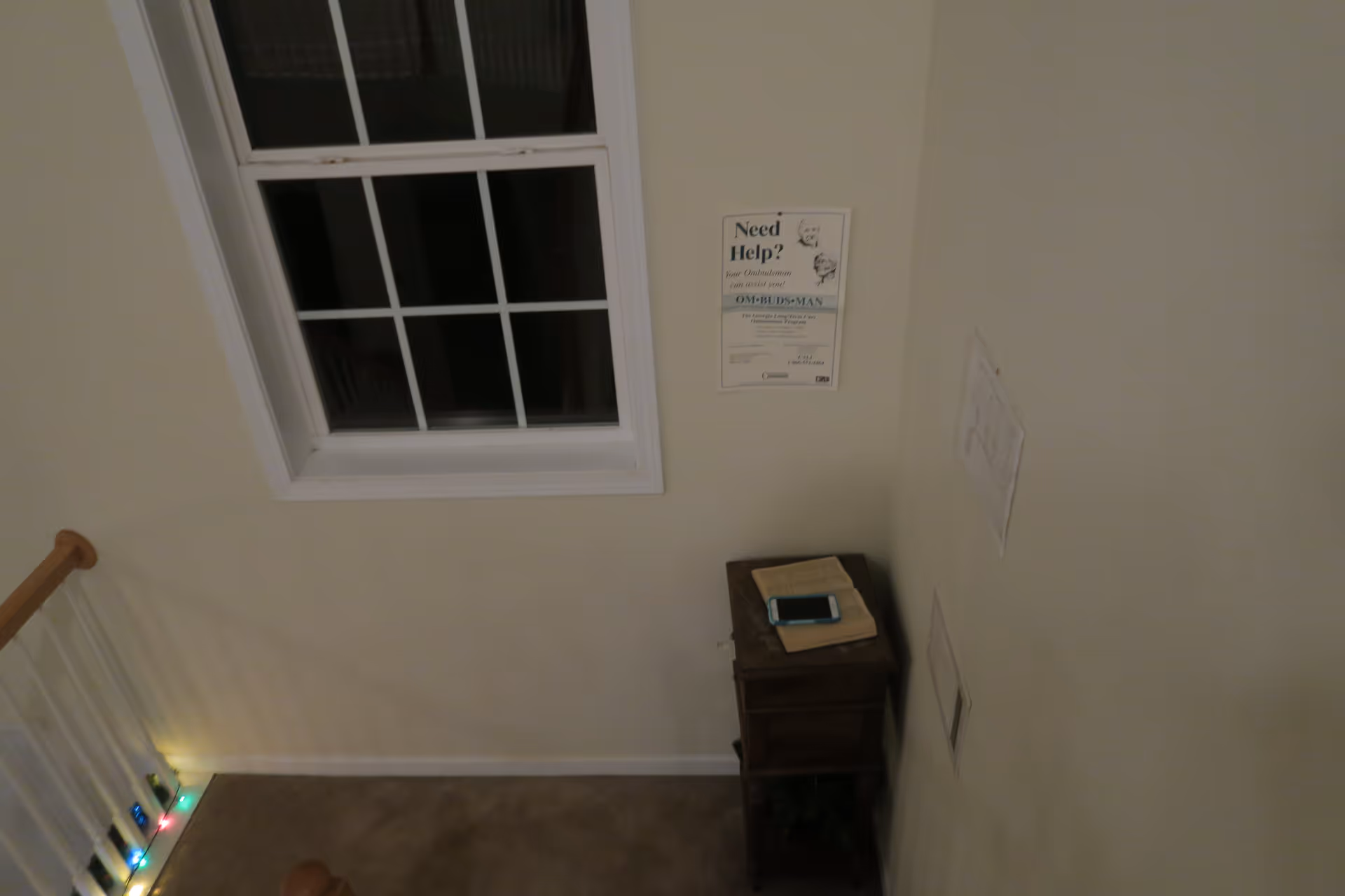 A small corner area with a window above a wooden side table that has a book and a smartphone on it. There is a poster on the wall that reads 'Need Help? Your Ombudsman can assist you!' and some other papers pinned to the adjacent wall. The floor is carpeted and there are colorful string lights along the stair railing.
