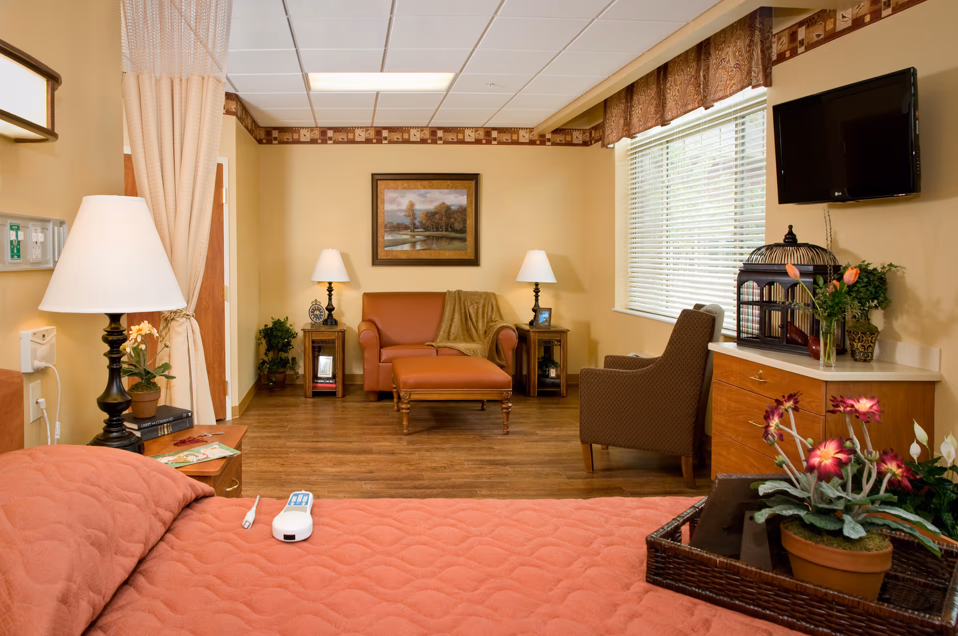 Cozy senior living suite with a bed in the foreground, a loveseat and armchair, lamps, TV, and decorative plants.