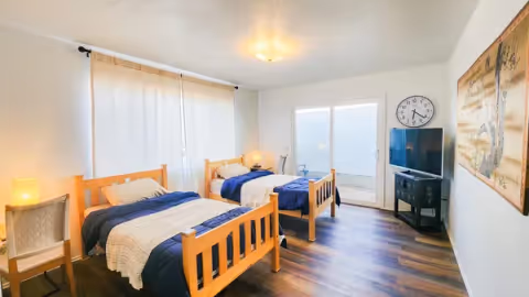 Bright bedroom with two wooden twin beds dressed in blue and white linens, a TV on a stand, a large wall clock, sliding glass door, and hardwood floors.