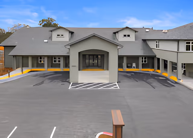 Front exterior view of a senior living facility building with a covered entrance, multiple windows, and a large paved parking area in front. The building is painted gray with a dark roof and surrounded by some trees in the background under a clear blue sky.