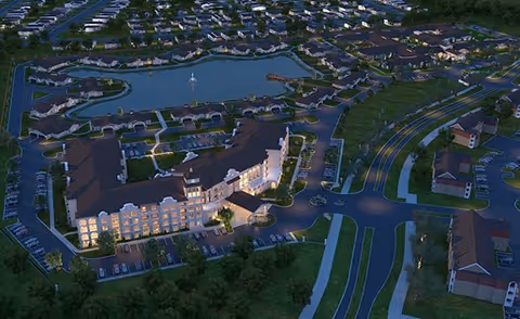 Aerial nighttime view of Wilshire Senior Living facility showing a large, well-lit main building surrounded by smaller buildings, parking areas, roads, and a central pond with pathways and landscaping.