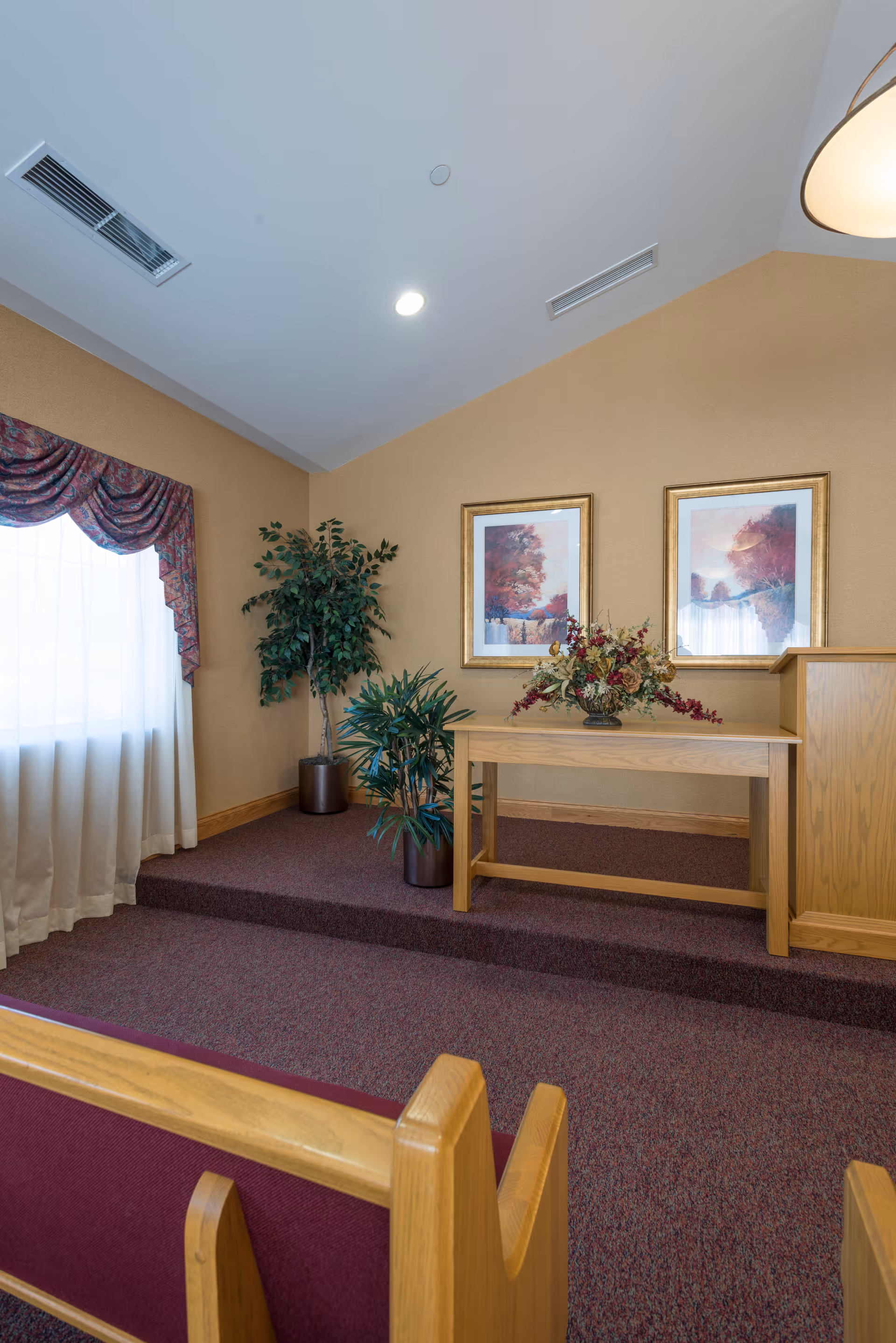 Interior of a room with a raised platform featuring a wooden table with a floral arrangement, two framed landscape paintings on the wall, two potted plants, and a window with sheer curtains and a decorative valance. Wooden pew-style seating with red cushions is visible in the foreground.