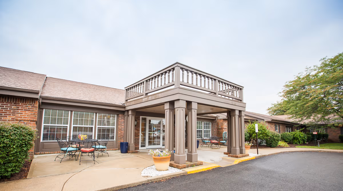 Covered entrance and portico of a brick senior living facility with outdoor seating, planters, and a driveway.