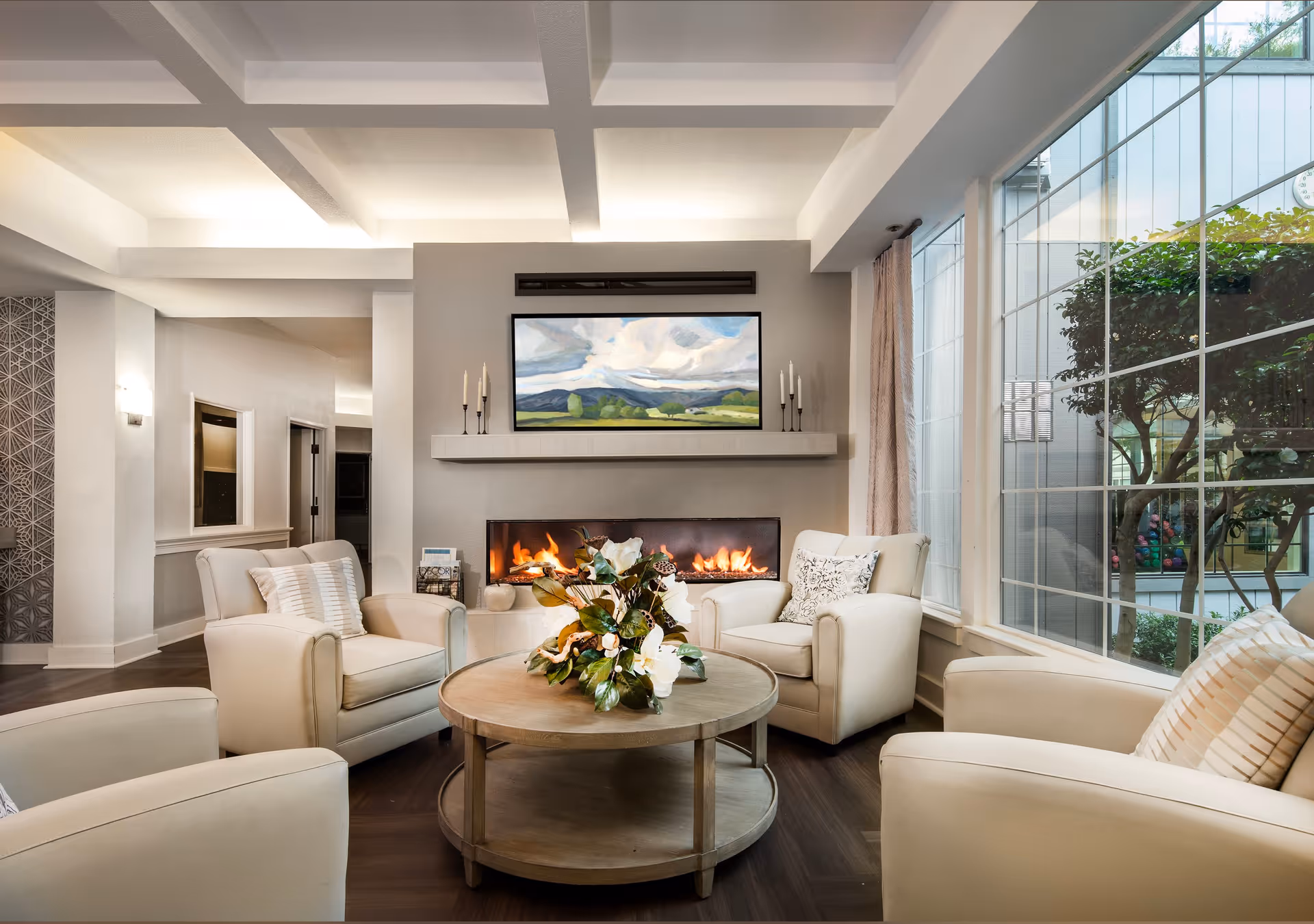 A cozy living room with four beige armchairs arranged around a round wooden coffee table with a floral centerpiece. A modern fireplace with a fire burning is set into the wall, above which hangs a landscape painting. Large windows on the right side let in natural light and show greenery outside.