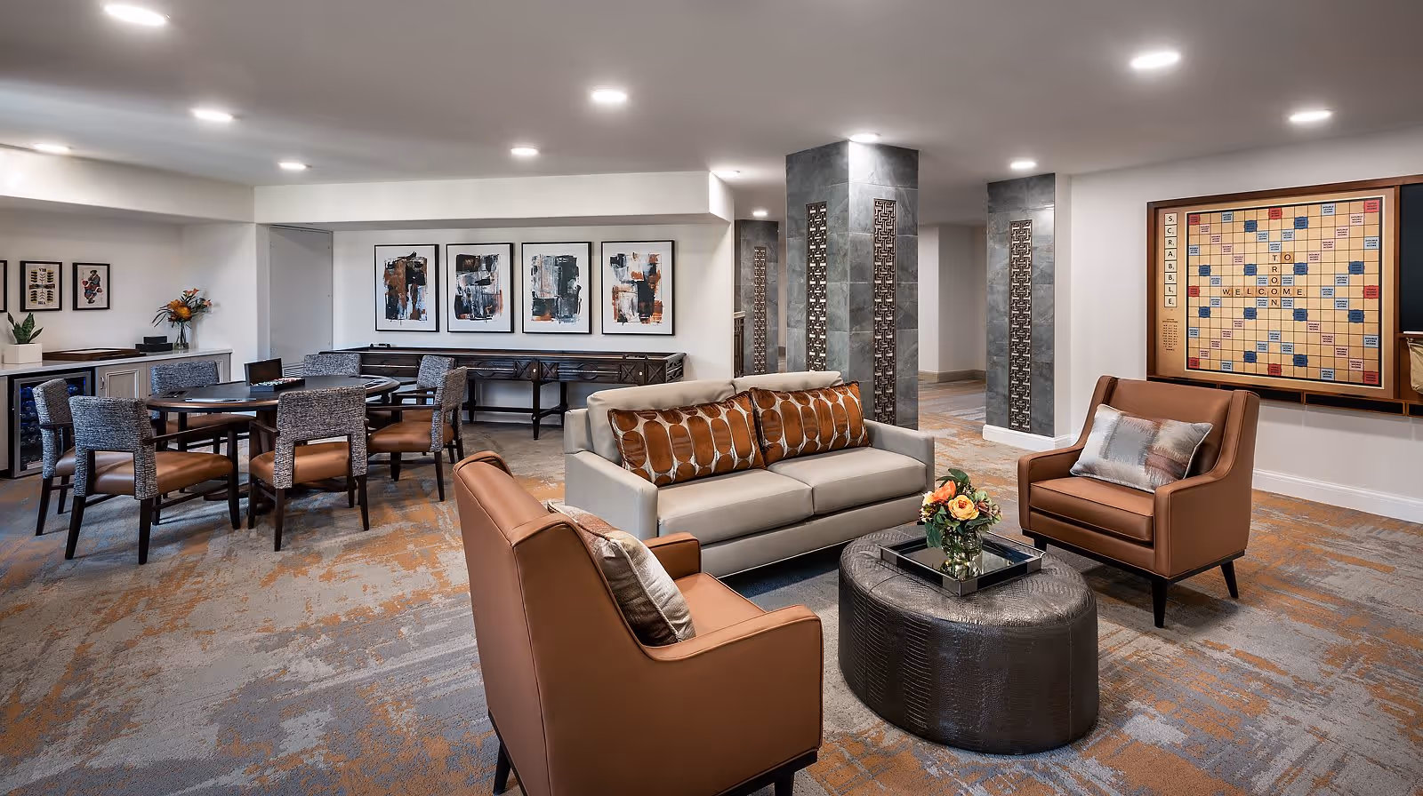 A cozy lounge area in a senior living facility featuring a beige sofa with patterned cushions, two brown leather armchairs, and a round black ottoman with a flower arrangement on top. In the background, there are several tables with chairs, abstract artwork on the wall, and a large Scrabble board mounted on the wall. The room has a carpeted floor and recessed ceiling lights.