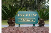 Green entrance sign reading "BAYVIEW Manor" surrounded by palm plants and mulch with trees in the background.