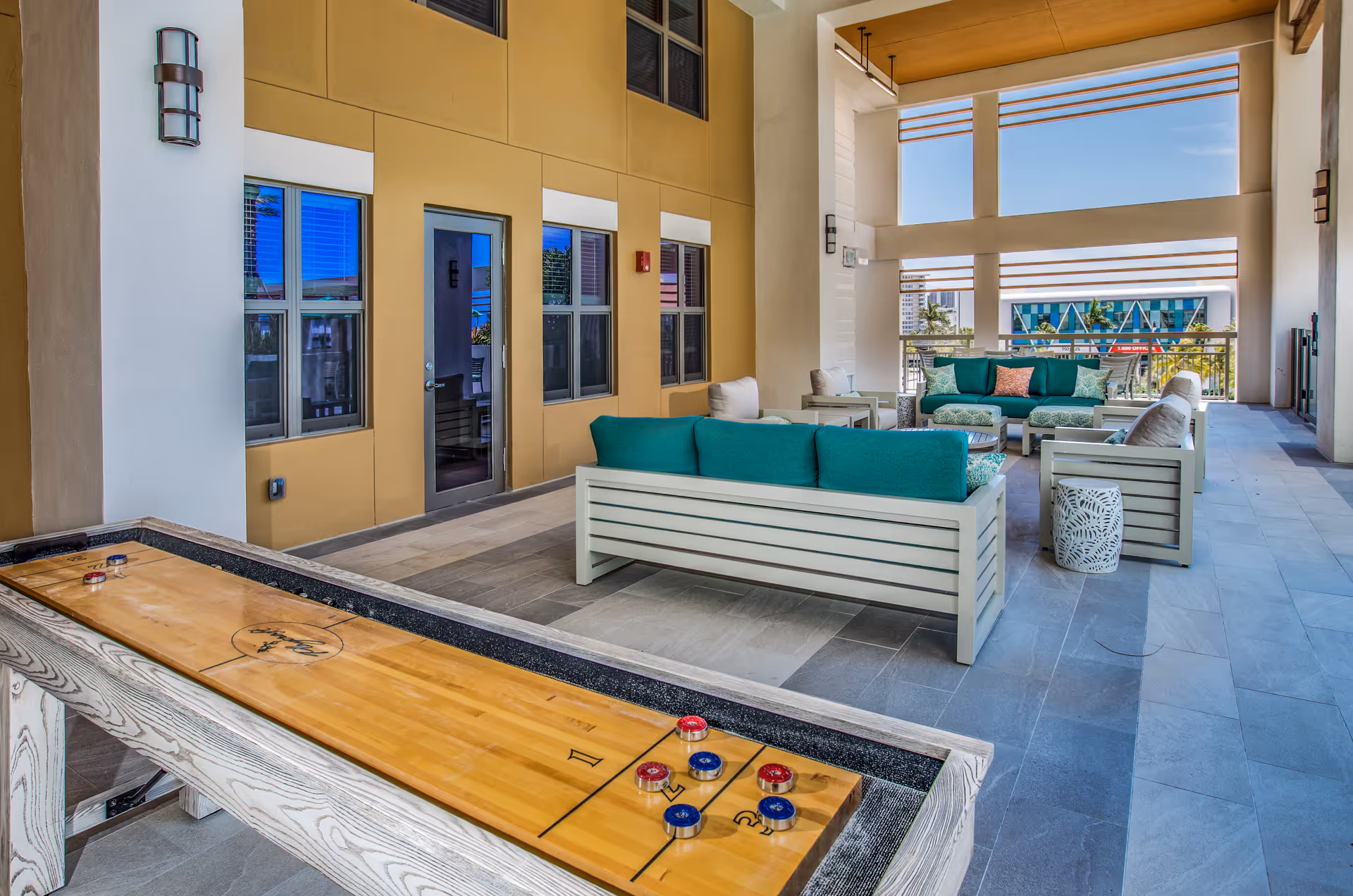 Covered outdoor lounge area with teal-cushioned sofas and a shuffleboard table.