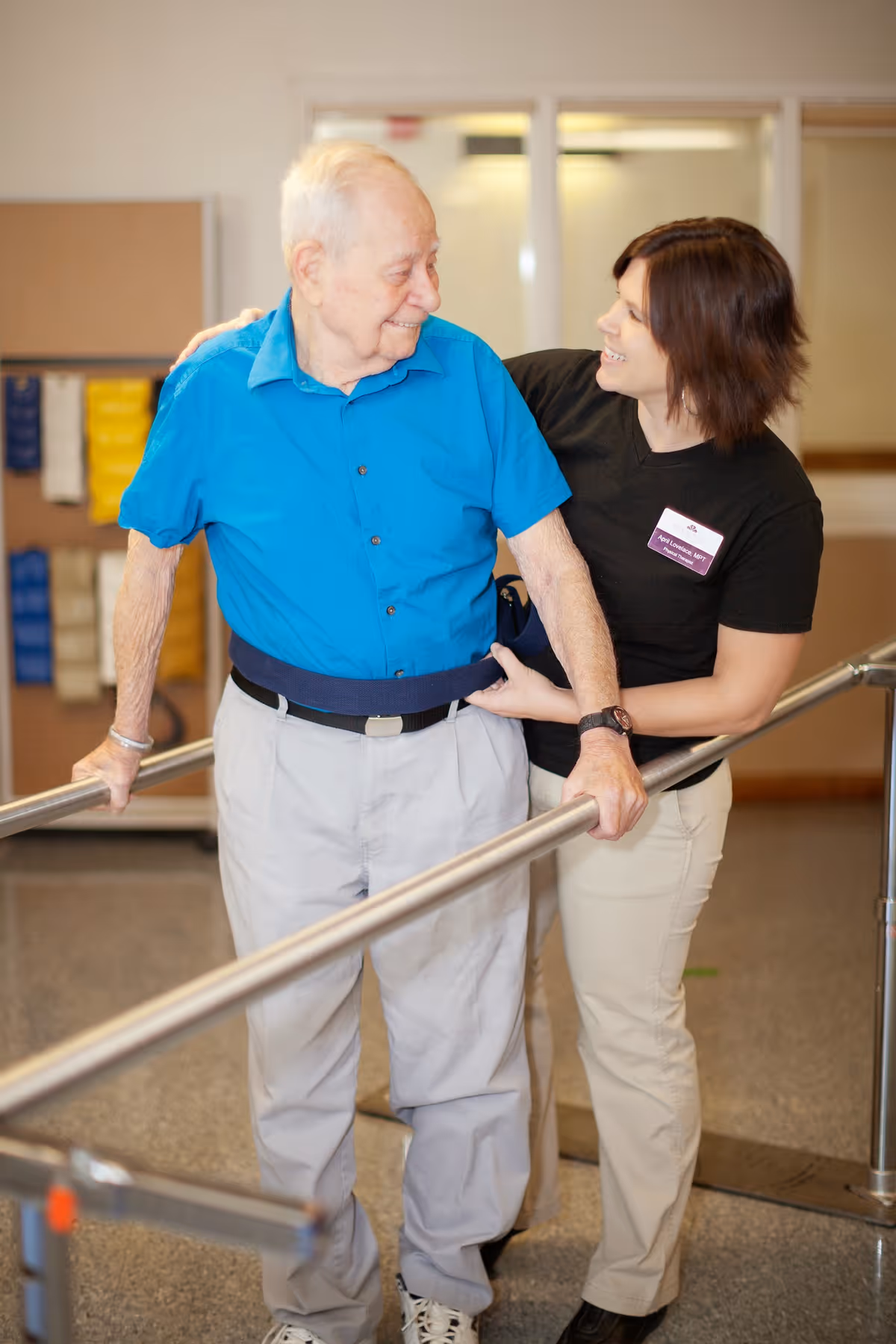 An elderly man in a blue shirt and light-colored pants is using parallel bars for walking support while a female caregiver in a black shirt and beige pants assists him, both smiling and looking at each other in a rehabilitation or therapy setting.