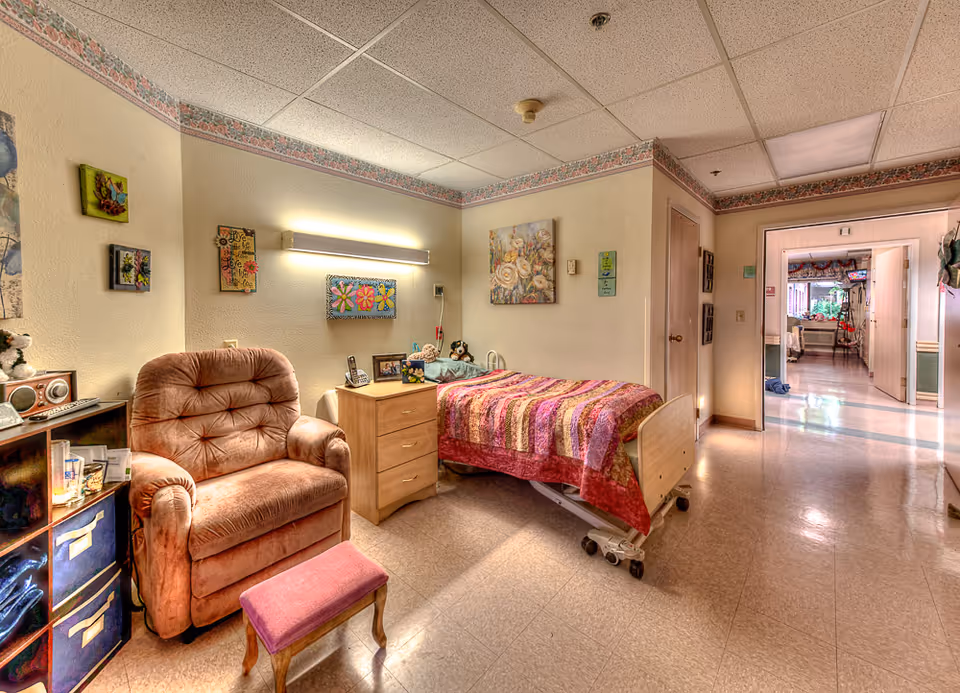 A cozy senior living room featuring a single hospital-style bed with a colorful quilt, a plush armchair with a small footstool, a wooden nightstand with framed photos and a telephone, and decorative wall art. The room has a tiled floor and a drop ceiling with fluorescent lighting. A hallway is visible in the background leading to other rooms.