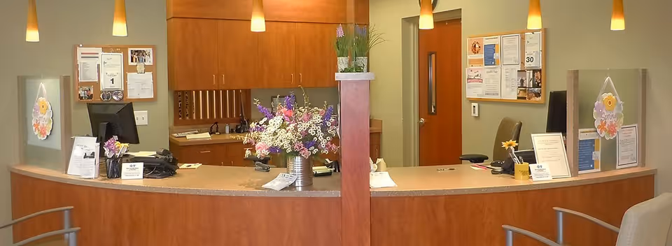 Reception desk area in a senior living facility with a curved wooden counter, two computer monitors, flower arrangements, bulletin boards with notices on the wall, and pendant lights hanging from the ceiling.
