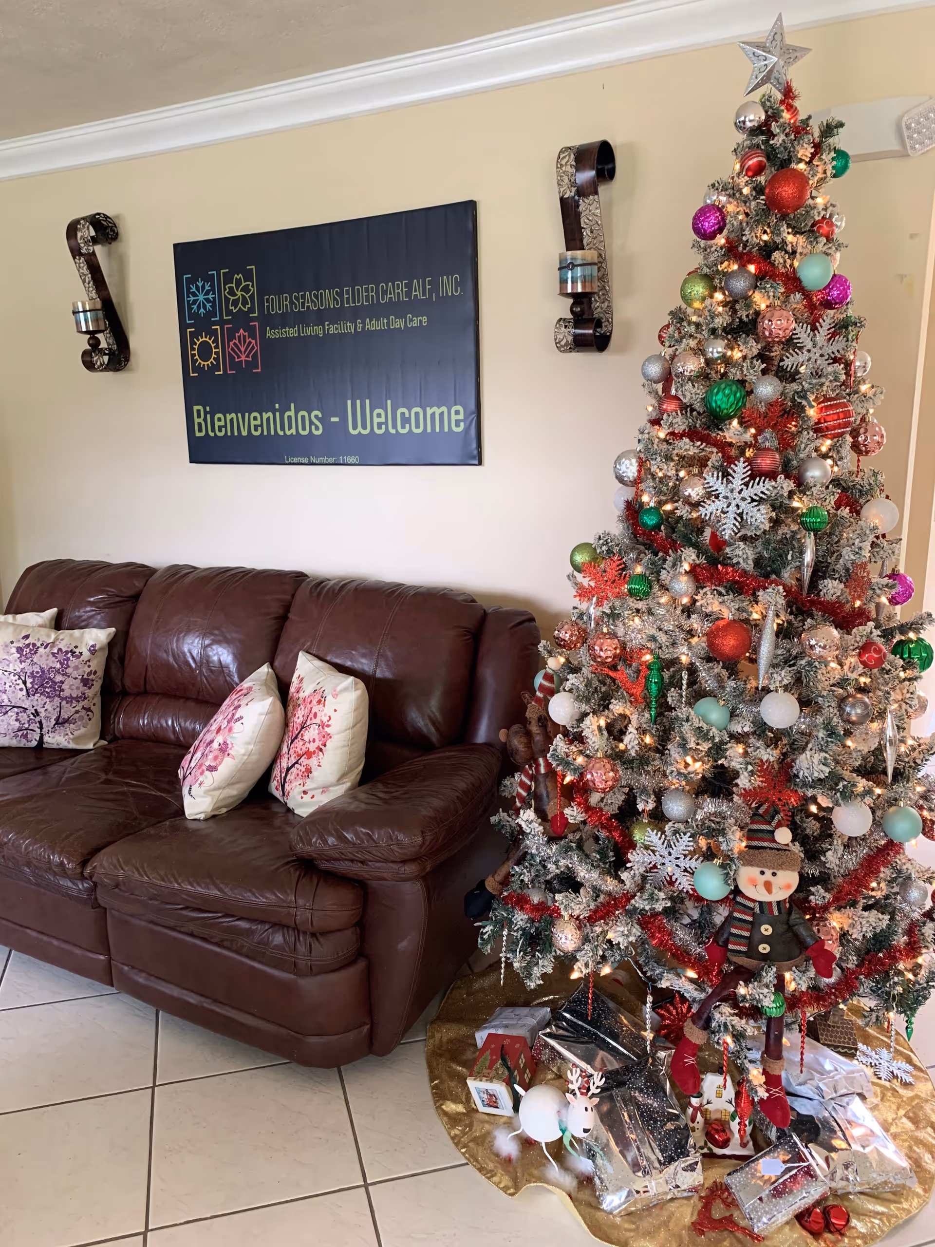 A cozy interior room with a brown leather couch adorned with floral pillows next to a decorated Christmas tree with ornaments, lights, and presents underneath. On the wall above the couch is a sign that reads 'FOUR SEASONS ELDER CARE ALF, INC. Assisted Living Facility & Adult Day Care Bienvenidos - Welcome'.
