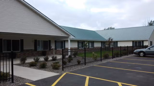 Exterior view of a single-story senior living facility building with a green metal roof, beige siding, and brick accents. There is a black metal fence enclosing a small landscaped area with shrubs and a concrete walkway. A parking lot with marked spaces is visible in the foreground under a cloudy sky.