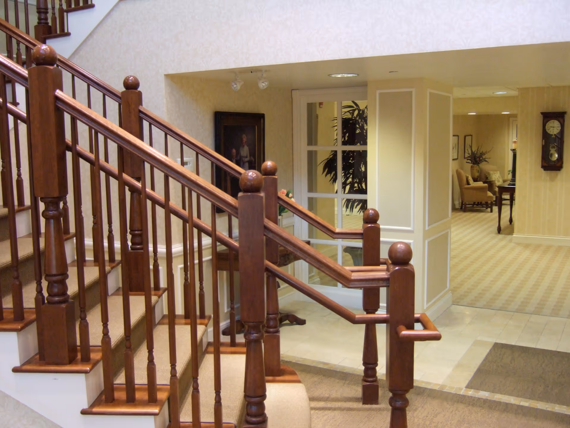 Interior view of a senior living facility showing a wooden staircase with carpeted steps and polished wooden handrails. In the background, there is a hallway with beige walls and carpet, a grandfather clock mounted on the wall, and a seating area with chairs and a table. A framed portrait and some plants are also visible.