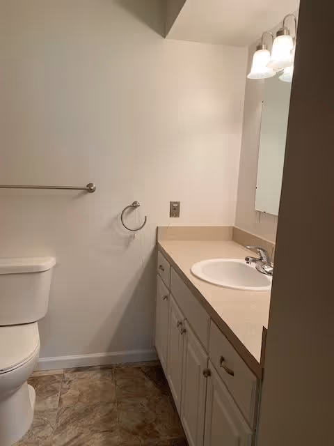 A small bathroom featuring a white toilet on the left, a beige countertop with a white sink and silver faucet on the right, white cabinetry below the countertop, a mirror above the sink, and a wall-mounted light fixture with three bulbs. The walls are painted white and the floor has brownish tile.