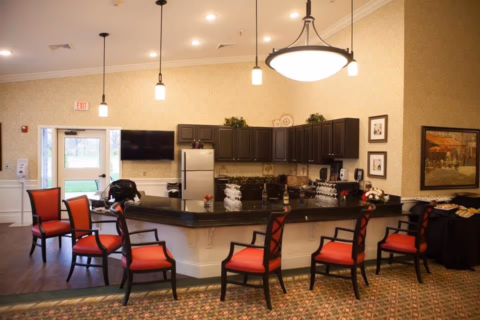 Interior view of a communal kitchen area with a large black granite countertop island surrounded by six red cushioned chairs. The kitchen has dark brown cabinets, a stainless steel refrigerator, and a mounted flat-screen TV on the wall. Pendant lights hang from the ceiling, and there is a door with a window leading outside. The room has patterned carpet and wallpapered walls with framed artwork and a table with refreshments on the right side.