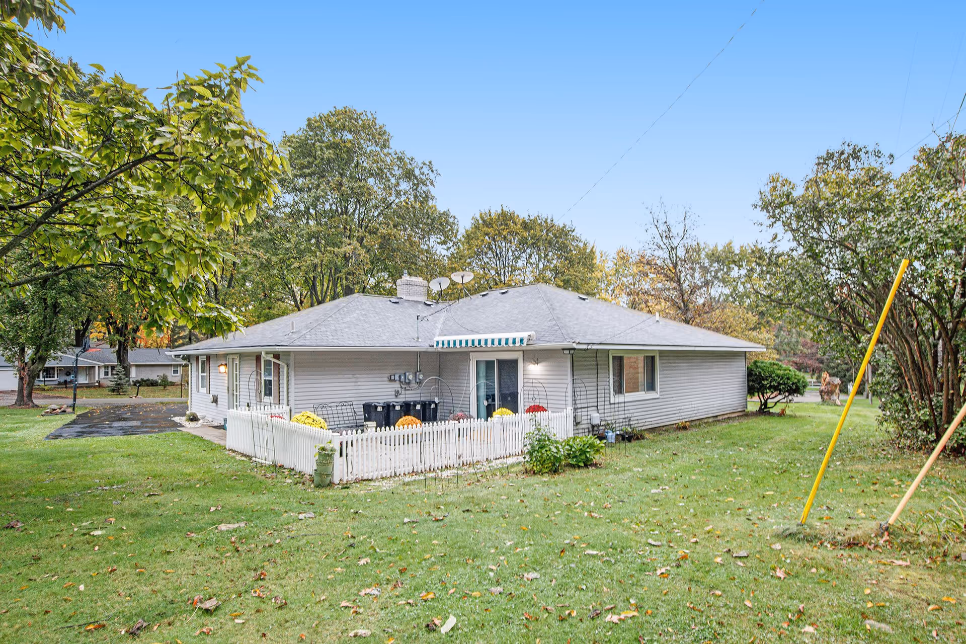 Single-story residential building with gray siding and a gray roof, surrounded by green grass and trees under a clear blue sky. There is a small white picket fence enclosing a patio area with colorful flowers and outdoor seating. Utility meters and trash bins are visible on the side of the building.