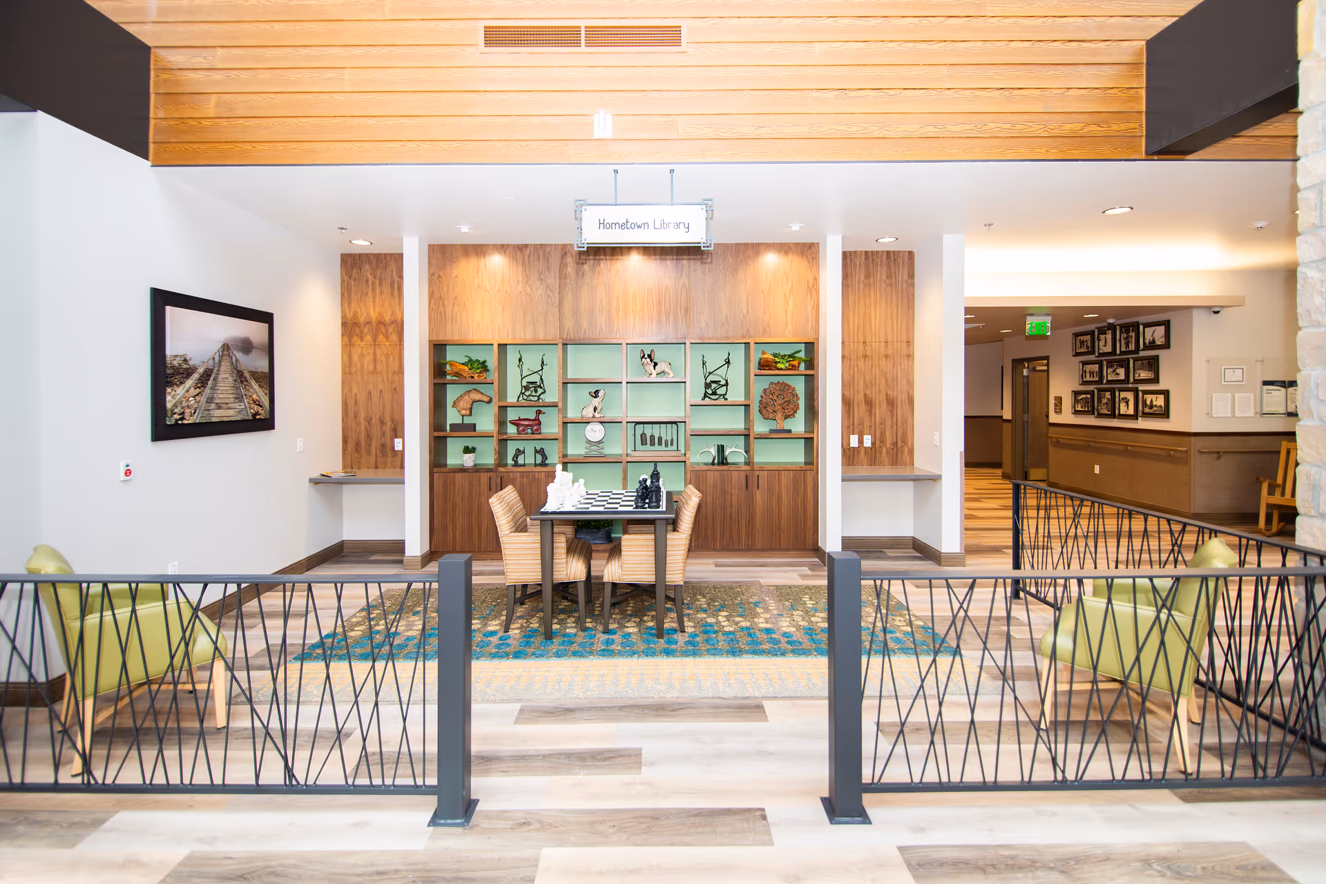 A bright and modern common area labeled 'Hometown Library' featuring a table with a chess set and four chairs. Behind the table is a wooden shelving unit with decorative items and art pieces. The space has light wood flooring, a colorful rug, two green armchairs separated by a black metal railing, and a hallway with framed pictures on the wall.