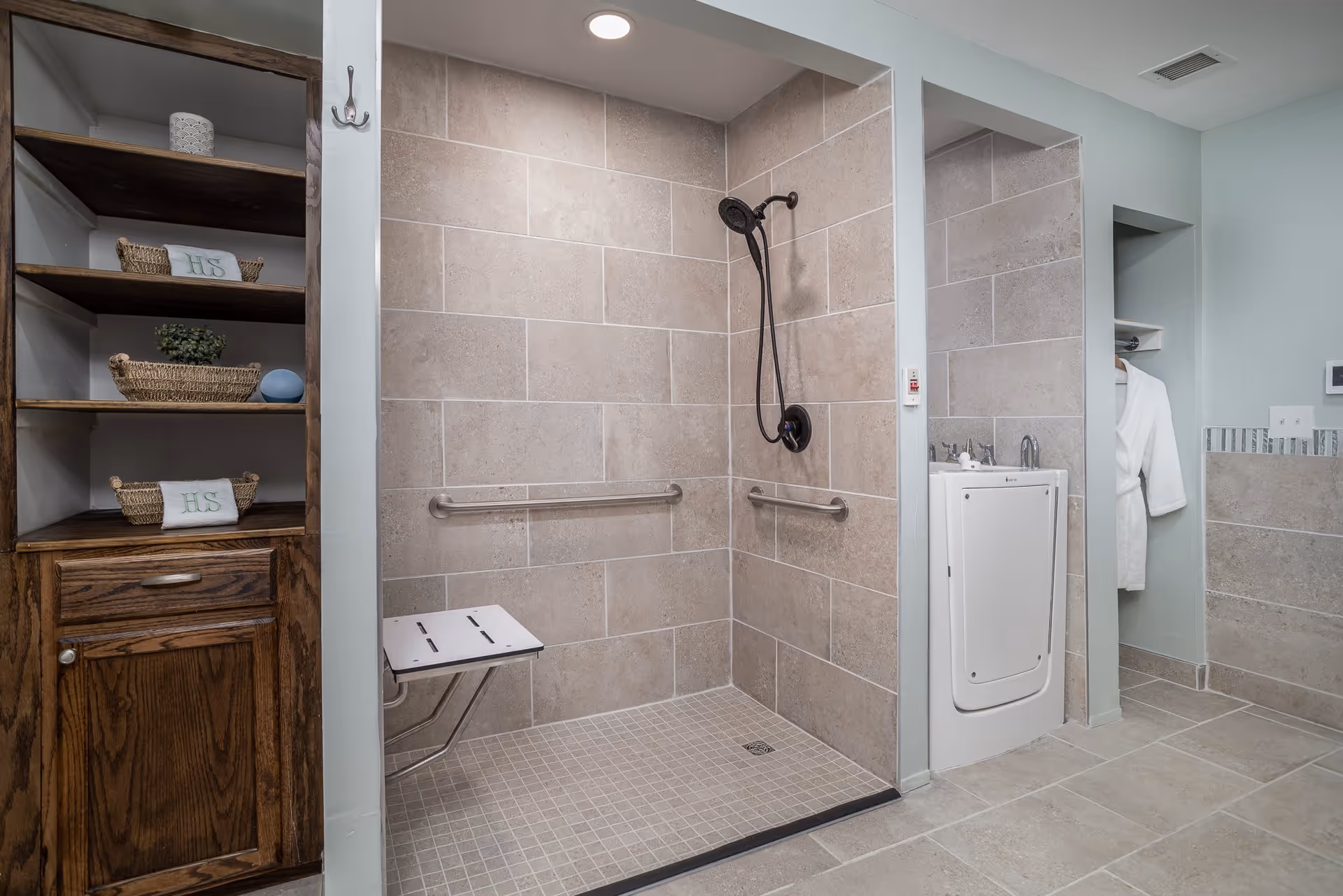A spacious, accessible bathroom featuring a walk-in tiled shower with a fold-down seat and grab bars. The shower has a handheld showerhead mounted on the wall. To the right of the shower is a white sink with a large mirror above it. On the left side, there is a wooden shelving unit with baskets and towels. The bathroom has light-colored tile flooring and walls, with a white bathrobe hanging in a small alcove.