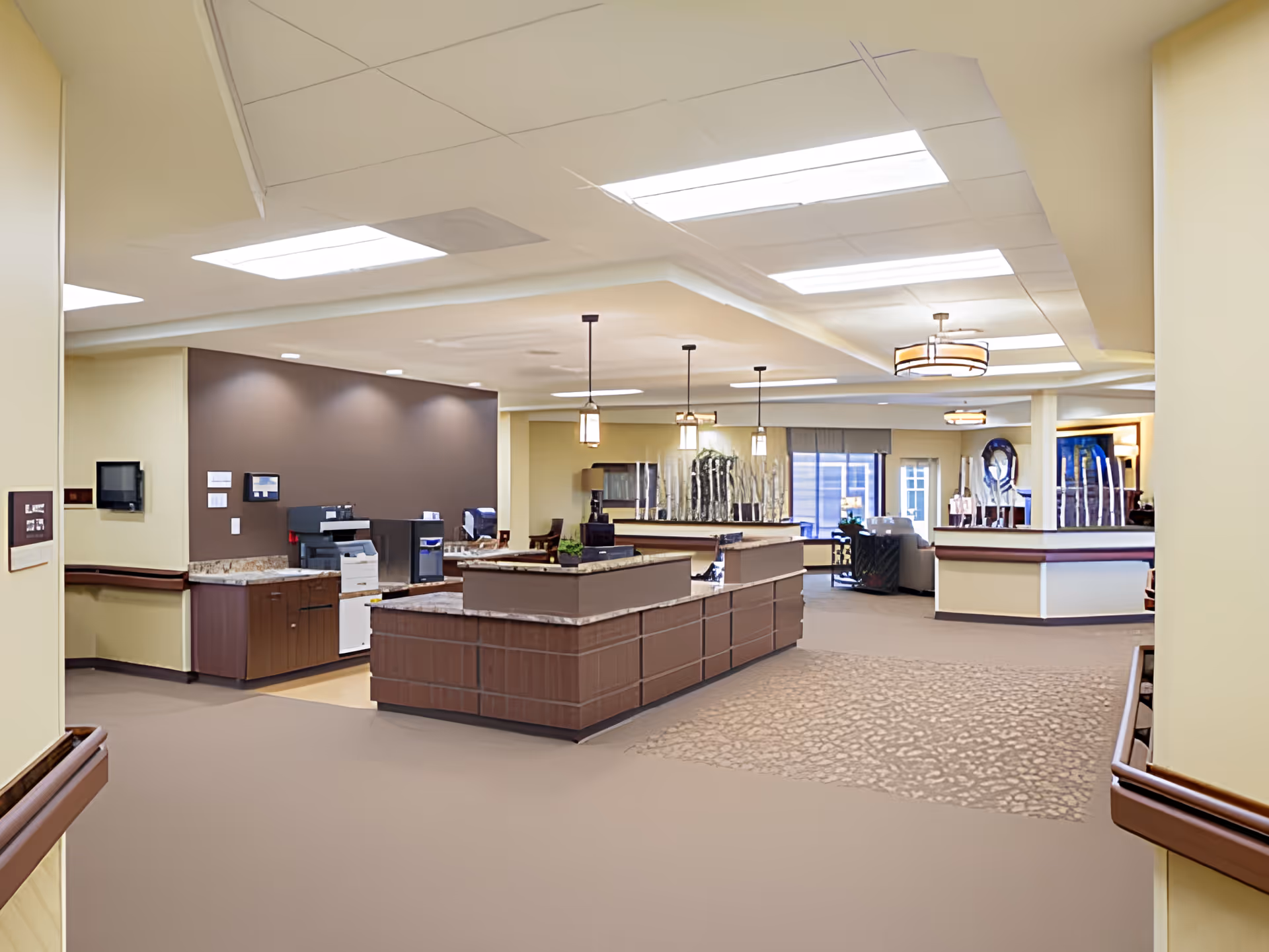 Interior view of a senior living facility reception area with a large wooden reception desk in the center, pendant lights hanging from the ceiling, and seating areas in the background. The walls are painted in neutral tones with a combination of beige and brown, and the floor has a mix of carpet and tile. The space is well-lit with ceiling lights and natural light coming through windows.