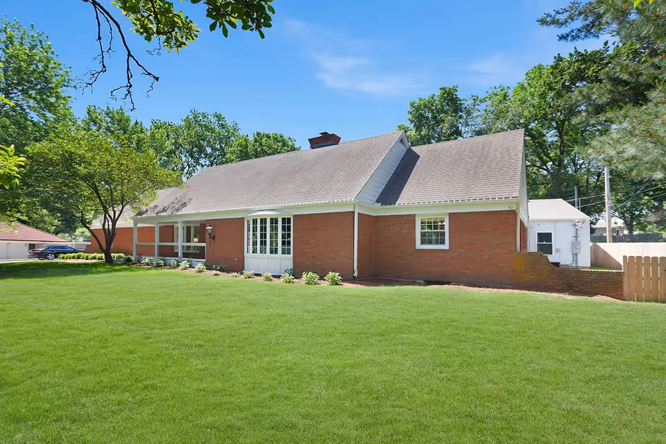 Single-story red brick house with a large green lawn, trees, and a clear blue sky.