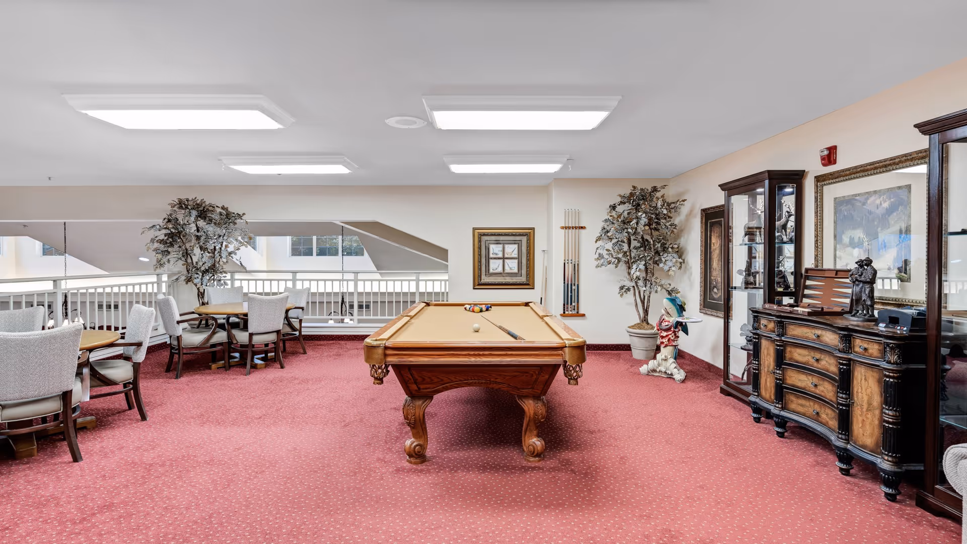 Carpeted communal game room featuring a billiards table centered, round seating tables, display cabinets and potted plants.
