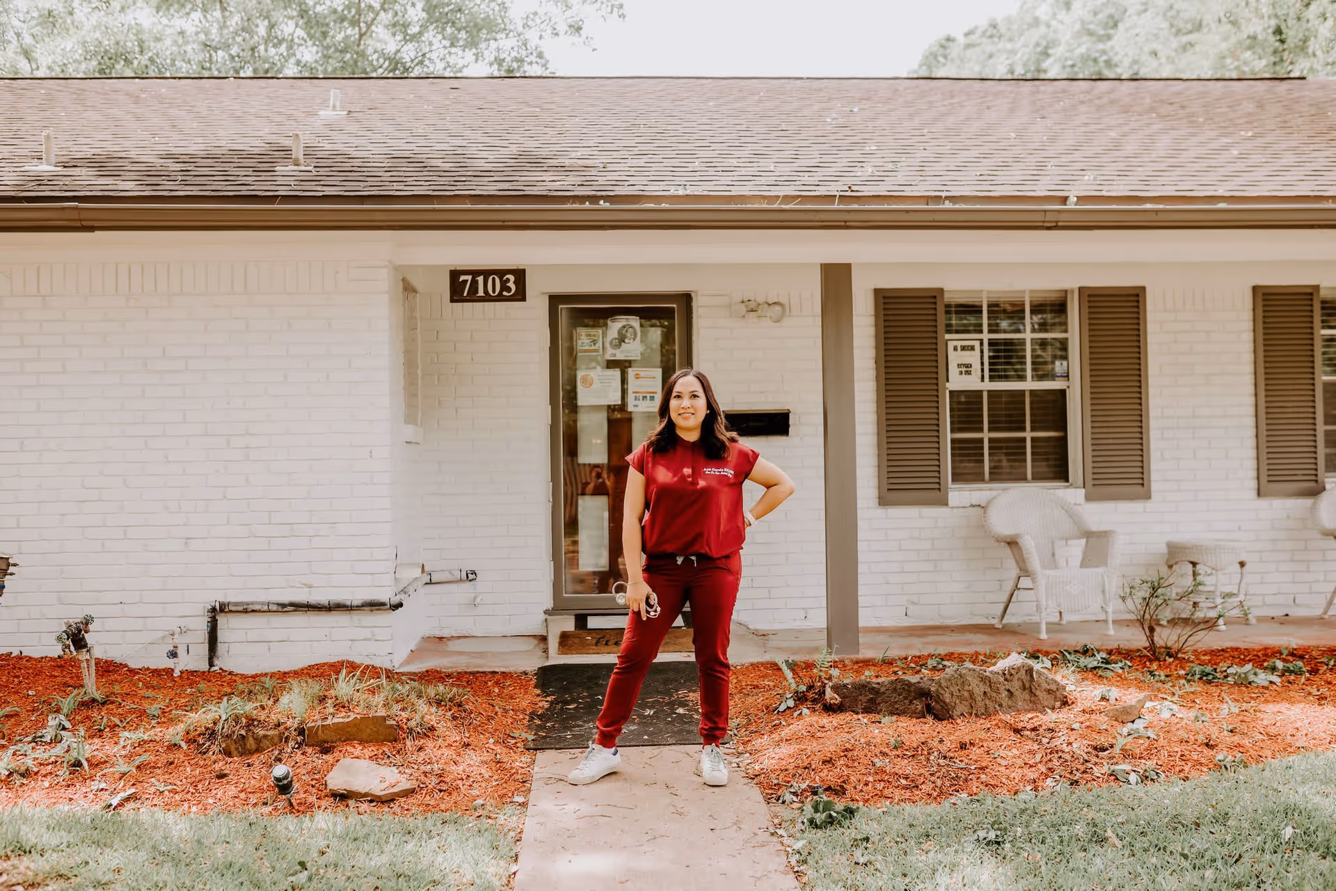 A woman wearing a red uniform stands confidently on the walkway in front of a single-story white brick building with the address number 7103. The building has brown shutters on the windows and white wicker chairs on the porch. There is a landscaped area with red mulch and some plants in front of the building.