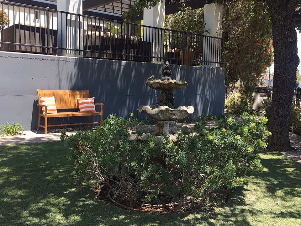 Outdoor courtyard with a tiered stone fountain surrounded by shrubs and a wooden bench with striped pillows against a gray wall.