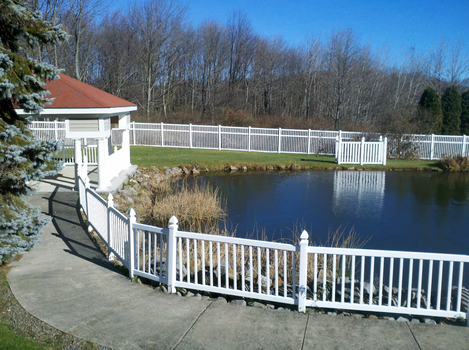 A peaceful outdoor scene featuring a small pond surrounded by a white picket fence. There is a paved walkway leading to a gazebo with a red roof on the left side. Trees without leaves are visible in the background under a clear blue sky.