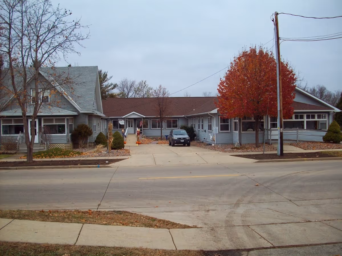 Exterior view of a single-story building with gray siding and a brown roof, featuring a driveway with a parked car and a tree with red autumn leaves near the entrance. The building appears to be a residential or care facility with a sidewalk and street in the foreground.